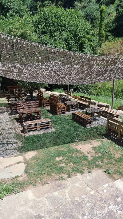The image depicts an outdoor seating area in Rupit, Spain, characterized by rustic, DIY furniture made from wooden pallets. The area is covered by a camouflage netting canopy, offering shade. The ground is a mixture of paved stones and grassy patches, with a backdrop of dense green foliage and trees, suggesting a natural, possibly wooded, environment. Several tables and benches, all constructed from pallets, are arranged for seating. Some of these are low tables with accompanying benches, while others are higher, resembling picnic tables. The furniture shows signs of wear and tear, contributing to the rustic aesthetic. There are no people visible in the scene. The overall impression is of a casual, environmentally conscious outdoor relaxation space.