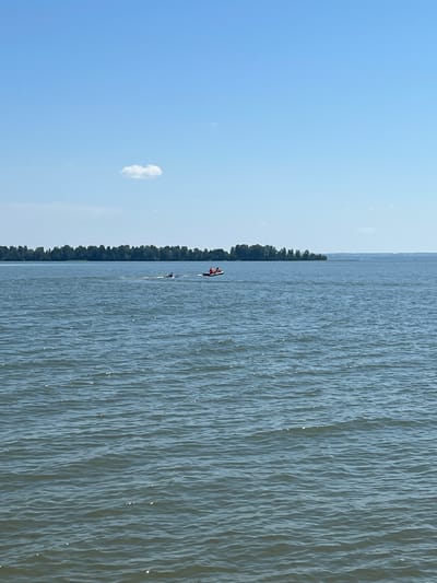 The image shows a wide, calm body of water under a clear blue sky with a few small clouds. In the distance, a dense line of trees forms the shoreline of a landmass. On the water, two jet skis are visible. The first jet ski, closer to the viewer, is being ridden by at least two individuals wearing life vests. A second jet ski is further to the right, also with at least one occupant. The water surface is rippled, indicating a gentle breeze. The overall scene is that of a recreational activity on a body of water, likely a lake or large river, on a bright, sunny day. No specific landmarks or urban features are discernible, making it impossible to ascertain a location within Borovoye, Russia.