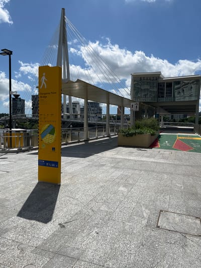 The image captures a vibrant, sunny day in a modern urban pedestrian area of Brisbane City, Australia. The central feature is the sleek, white cable-stayed Kangaroo Point Bridge, which dominates the left side of the frame with its elegant pylon and numerous tensioned cables stretching into a bright blue sky dotted with fluffy white cumulus clouds.

In the foreground, a prominent yellow information sign stands vertically on the paved walkway. The sign clearly displays the text "Kangaroo Point Bridge" along with a pedestrian symbol and a map of the surrounding area, indicating the bridge spans a body of water, likely the Brisbane River. The ground is paved with light grey, textured rectangular tiles, reflecting the strong sunlight. Shadows cast by the sign and nearby structures suggest it is mid-morning or early afternoon.

Modern multi-story buildings with reflective glass facades are visible in the background, appearing both behind and beneath the bridge's structure. A small planter box with green foliage adds a touch of nature near one of these buildings. A white sign with symbols for both pedestrians and cyclists is visible further along the path, indicating shared usage. No people are visible, giving the scene a serene, open feel, but the infrastructure suggests it is a popular route for walking and cycling. The overall impression is one of a clean, well-maintained, and visually striking urban landscape.