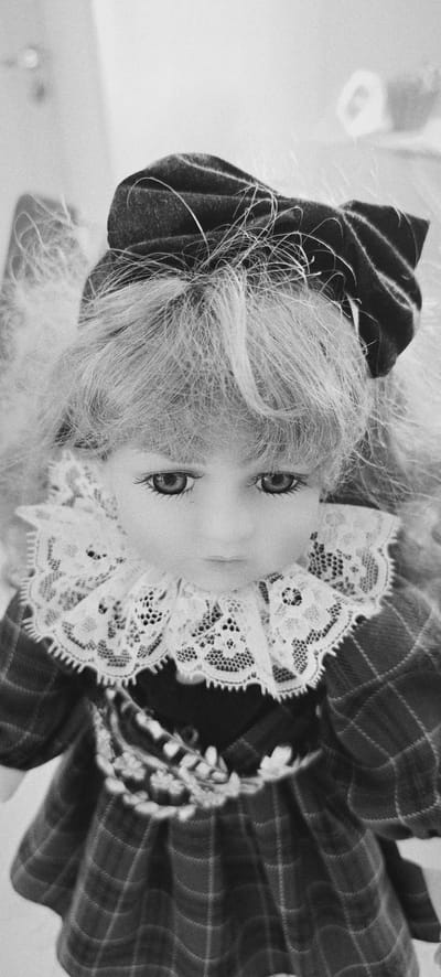 A close-up, black and white, eye-level, portrait shot shows a doll's face. The doll has pale skin, blue eyes with dark lashes, and blonde hair. She is wearing a dark bow on her head and a white lace collar. Her dress is plaid. The background is blurred and appears to be an indoor setting. The doll is looking down. The image is grainy and has a vintage feel.