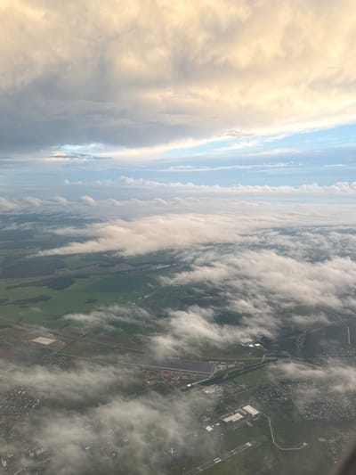 This aerial view, taken from an airplane, shows a landscape partially obscured by clouds. The scene features a patchwork of green fields, some with agricultural divisions, interspersed with areas of dense woodland. Roads are visible, connecting scattered settlements and industrial or commercial buildings. One prominent feature is a large, rectangular building complex with a white roof, likely an industrial warehouse or logistics center, and a runway with markings indicating an airport. In the distance, smaller towns and villages with residential buildings are visible. The sky above is a mix of soft clouds illuminated by sunlight, creating a gentle gradient from yellow-orange near the horizon to blue overhead. A faint outline of another airplane can be seen on the ground at the bottom right of the image. Based on visual cues such as the agricultural patterns, presence of an airport, and architectural styles, this scene is consistent with the outskirts of the city of Aramil, Russia, near the Koltsovo International Airport.