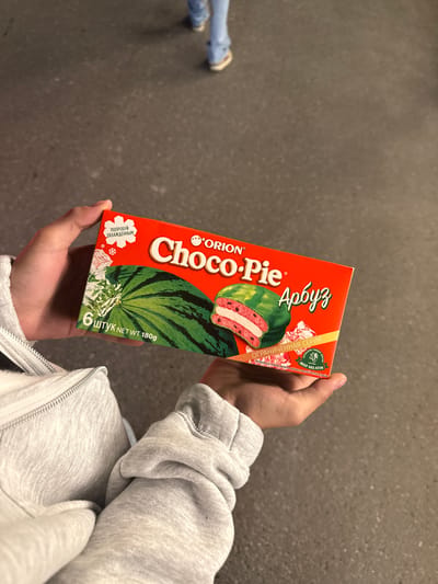 A person wearing a light grey zip-up hoodie is holding a box of "Orion Choco-Pie Арбуз" (Watermelon) snacks. The box is red with images of watermelon and the product itself, and text in both English and Russian. The person's other arm is visible, also wearing the same hoodie. In the background is a grey surface, possibly a sidewalk or floor. Parts of another person's legs, clad in jeans and sneakers, are visible in the background, to the left.
The location is unspecified, and there is nothing visible that indicates the city of Tsakhkadzor, Armenia.