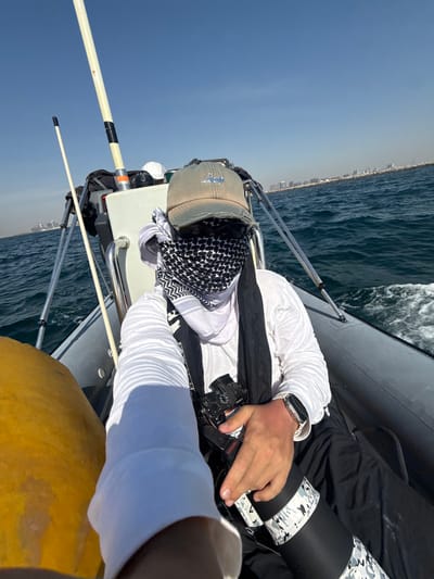 A man is on a boat at sea. He is wearing a black and white keffiyeh, a white long-sleeved shirt, and a silver Apple Watch. He is holding a black camera. The boat has gray inflatable sides and metal railings. The sea is choppy. In the background, a city skyline can be seen. This is likely Deira, United Arab Emirates. A yellow cushion is visible in the foreground.