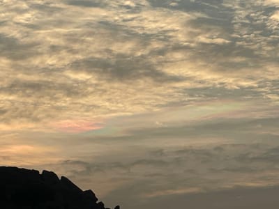 The image displays an expansive sky over Sanapur, India, dominated by layered cloud formations. The clouds, appearing to be altocumulus, exhibit a textured pattern of ripples and patches, predominantly illuminated with a warm golden-beige hue in the upper portions, gradually shifting to a grayish tone in the mid and lower sections. A distinct area of cloud iridescence is visible horizontally across the mid-left to mid-right of the frame, presenting soft pink, green, and light blue spectral colors within the cloud layers. A dark, silhouetted formation of jagged rocks occupies the lower left quarter of the image, contrasting sharply with the illuminated sky. There are no discernible people, animals, man-made structures, or specific actions or interactions depicted within the scene.