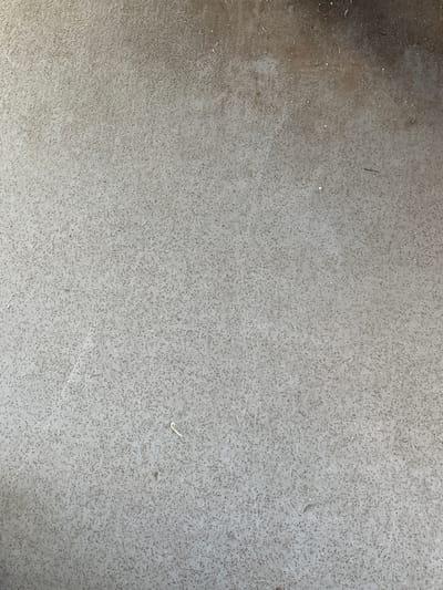 The image shows a close-up, overhead view of a mottled, light gray concrete surface. The surface is textured with numerous small, dark specks and occasional lighter flecks, giving it a speckled appearance. There are a few very small, light-colored debris particles scattered across the surface. In the upper right quadrant, the surface appears darker and possibly wet or stained. A thin, curved, light-colored fiber or strand is visible near the bottom center of the image. The lighting suggests an outdoor environment, possibly overcast or in shadow. The context of Hlevakha, Ukraine is not visually present in the image.
