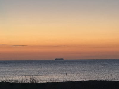 This media file depicts a serene seascape at dusk. The subjects are a large cargo ship silhouetted on the horizon and the calm, rippling water of the sea. In the foreground, the dark silhouette of beach grass and sand dunes frames the scene, suggesting a coastal location. The sky displays a gradient of soft colors, transitioning from a pale gray at the top to a warm orange hue near the horizon, indicative of either sunrise or sunset.
There are no people visible in the image. The activity taking place is passive; the cargo ship appears to be stationary or moving slowly, and the sea is relatively calm. The time of day is clearly dusk or dawn due to the sky's coloration. The weather appears to be clear with a few faint clouds visible in the distance. The location is stated to be Thanet, United Kingdom, which aligns with the coastal seascape. The overall mood evoked is one of tranquility and peacefulness. No text is visible in the media file.
