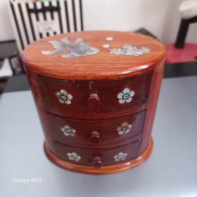 The image shows a wooden jewelry box with three drawers. The top of the box is round and features a decorative inlay of a fish and some coral. The drawers have small knobs and are adorned with inlaid flowers. The jewelry box is placed on a gray surface. In the background, there is a blurred view of vertical black and white stripes. The image also contains the text "Galaxy M32" in the lower left corner. The location is not discernible from the image.