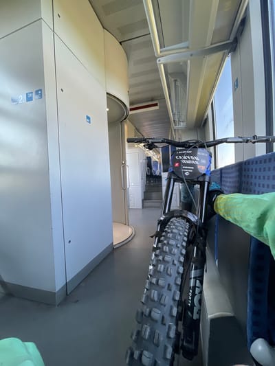 The image shows a close-up, eye-level, indoor shot of a downhill mountain bike parked inside a train car. The bike's front wheel and suspension are prominent in the foreground, with the handlebars and front number plate visible. The number plate displays "CANYON ENDURO" and other branding like "SRAM" and "FOX". A person's legs are visible on the right, wearing a green jacket with yellow speckles and blue gloves. The background reveals the interior of a train, with seats visible further down the aisle and doors on the left wall. There are signs indicating "WC" and "3M" on the wall. The overall environment suggests a mode of transport, likely a train, designed for carrying passengers and possibly equipment. The image does not contain any information about the city of Merichleri, Bulgaria.