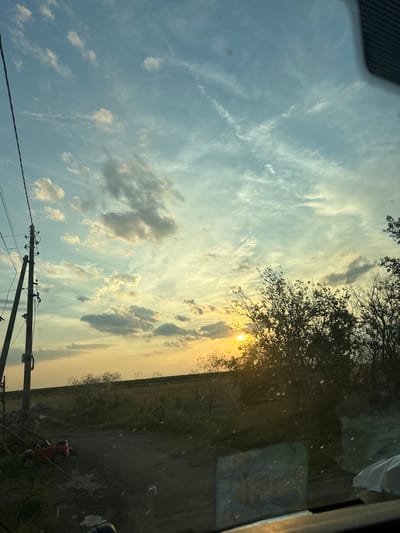 The image depicts a rural sunset scene in Mlekarevo, Bulgaria. A power line pole stands to the left, with visible wires and insulators. In the foreground, a small red toy car is parked on a gravel path. Beyond the path, a field stretches towards the horizon, bordered by trees and bushes. The sky is filled with clouds of varying shades of blue, gray, and yellow, illuminated by the setting sun, which is partially obscured by foliage in the mid-right of the frame. The overall impression is one of natural beauty and a serene, quiet atmosphere.