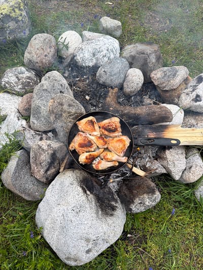 The image depicts an outdoor cooking scene amidst a rocky, grassy environment, possibly in the vicinity of Mazeri, Georgia. A black frying pan filled with pieces of cooked chicken is placed on a makeshift campfire constructed from large stones and logs. Smoke rises from the fire, indicating it is still active. The chicken pieces appear browned and well-cooked. The surrounding area is covered in green grass and scattered small purple flowers. The rocks are of various sizes and textures, some appearing to be granite. The overall impression is one of rustic, outdoor food preparation. No people are visible in the frame.