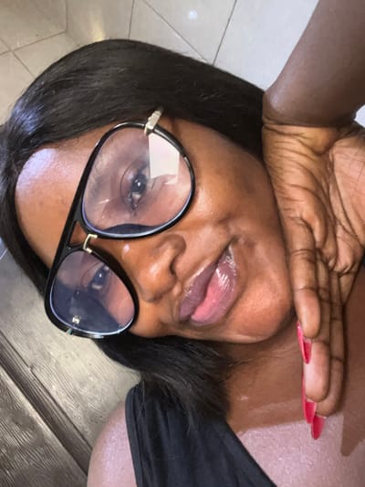 A diagonally-oriented close-up portrait shows an individual of dark complexion, located in Dzhidu, Nigeria. The subject wears black-framed aviator-style eyeglasses with reflective clear lenses. Dark, straight hair frames the face. The mouth is slightly open, revealing the tip of the tongue and glossy lips. The left hand, adorned with long, pointed red fingernails, is positioned against the left side of the face and neck. A dark-colored garment, possibly black, is visible on the upper torso. The background consists of a light-colored tiled surface in the upper right and a dark, wood-patterned surface in the lower left.