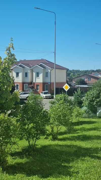 A sunny day with a clear blue sky is depicted in this image, likely taken in the city of Верзилово, Russia, given the context. The foreground is dominated by lush green grass and trees, with shadows cast by the foliage. In the midground, a street is visible with two cars parked. One is a silver SUV, and the other is a silver sedan. Behind the street, a modern two-story duplex-style house with a brown roof and cream-colored walls stands prominently. The house has multiple windows and a brick lower level. A yellow diamond-shaped road sign is positioned to the right of the house. In the background, more houses and a line of trees can be seen against the horizon. A tall street lamp is also visible on the left side of the image.