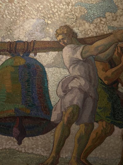 This is a detail from a fresco painting depicting two muscular men carrying a large bell. The man in the foreground, with blond hair and wearing a light-colored tunic, strains to lift a thick wooden beam supporting the bell. His body is angled forward, emphasizing his exertion. Behind him, another man, partially obscured, wears a green garment and also appears to be contributing to the effort. The bell itself is massive and colored in shades of green, blue, and dark brown. The background is textured and abstract, with hints of white and light blue suggesting a sky or clouds. The style is impressionistic, with visible brushstrokes creating a dynamic and textured surface. This fresco is located in the interior of the Santiago de Compostela Cathedral in Spain, specifically in the Ascension Chapel.