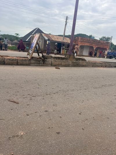 In Kona Garu, Nigeria, a street scene unfolds under an overcast sky. A dirt road runs alongside a concrete barrier. In the background, several structures are visible, including a building with a corrugated metal roof and an orange-colored building with arched doorways. A purple lamppost stands prominently in the foreground.
Several people are present. On the left, a man in a white shirt and dark pants stands near a sign. To his left, another man in a brown shirt and dark pants is visible. A woman in a burgundy garment walks by a child in a light-colored outfit.
In front of the corrugated metal building, a wooden easel holds a sign with a picture of a person and text. A woman in a colorful patterned dress stands near a young child. Further to the right, a man in a brown shirt stands outside the orange building, near another person in red. A blue truck with "AGRO" written on its side is parked to the far right. The ground is paved with asphalt and has some debris.