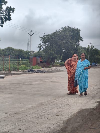 Two women, one in an orange patterned sari and the other in a blue patterned sari, are walking on a paved surface in Walsawangi, India. The woman in orange is gesturing towards her head. The background includes trees, a chain-link fence, a building with a red corrugated metal side, a pile of dark earth, and power lines against an overcast sky.