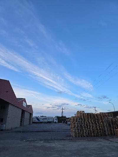 The image displays an outdoor industrial or commercial yard in Valverde del Majano, Spain, under a blue sky exhibiting scattered, elongated white clouds and a faint aircraft contrail. Overhead power lines are visible in the upper right. The foreground features a paved surface, segmented by a chain-link fence running horizontally across the lower third. To the left, a building with light-colored, possibly concrete, walls and a distinctive reddish-brown, corrugated, multi-gabled roof is present. Behind the fence, a white semi-trailer truck with blue branding is parked near the building, with a second white truck partially visible further back. To the right, a large, rectangular stack composed of numerous light-colored wooden pallets is prominently situated. In the background, behind the pallet stack, a utility pole with multiple electrical wires is visible, along with distant green foliage and the top of a curved street light. No individuals are depicted in the scene.