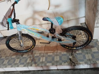 The image shows a light blue BMX bicycle with a turquoise seat and grips, and a black frame. The bicycle is wrapped in plastic, suggesting it is new or being protected. The word "MORTEIN" is visible on the tire rim. The bicycle is on a tiled floor that appears to be outdoors, possibly in a courtyard or paved area. The background consists of a tiled wall. Based on the tiling style, the presence of what appears to be a concrete wall with some discoloration, and the general outdoor setting, this scene is consistent with a location in Pekanbaru, Indonesia, specifically in a residential or commercial outdoor area. No people are visible in the image.