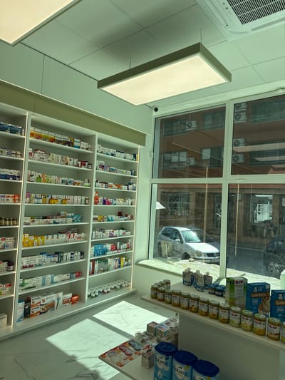 The image shows the interior of a pharmacy in Madan, Bulgaria. The shelves are stocked with a variety of products, including baby food, formula, and medicine. The products are neatly arranged, and the lighting is bright and clean. Through the window, the street outside can be seen, with a car and buildings. The overall impression is one of a well-organized and modern pharmacy. Visible text includes brand names like HIPP, Aptamil, and Gerber, as well as product names like "Gastrova" and "Bestiflex". The time of day appears to be daytime, and there are no visible weather conditions. There are no people visible in the image, but the presence of products suggests a commercial setting.