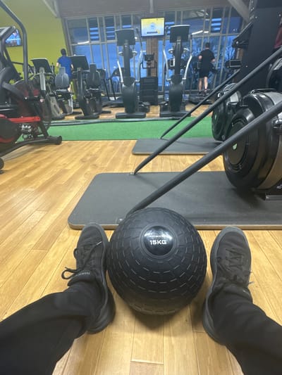 The image shows an indoor view of a fitness center in Welshpool, United Kingdom. The foreground is dominated by a 15kg black medicine ball, flanked by two feet clad in black athletic shoes and black pants. Adjacent to this setup is a rowing machine, with its dark components visible. The floor is constructed of wood panels. In the background, multiple exercise machines are arranged, and individuals are engaged in physical activity. Through a large window, a glimpse of the exterior landscape is visible, including trees and the twilight sky.