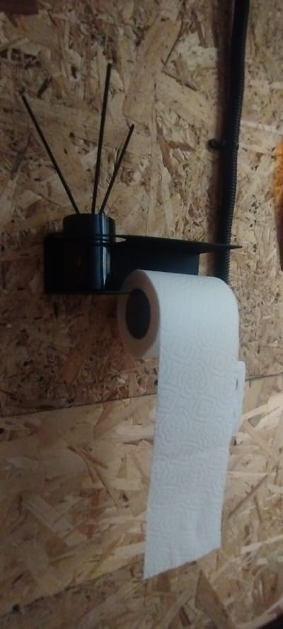 A close-up shot shows a roll of toilet paper hanging from a black holder attached to a wall. The wall appears to be made of oriented strand board (OSB), a wood-based material commonly used in construction. The toilet paper roll is white and has a textured pattern. To the left of the toilet paper holder, there is a black cylindrical object with several thin black sticks protruding from it, resembling an air freshener. The scene is indoors, likely in a bathroom or utility area, with a black conduit running vertically along the wall in the upper right portion of the image. The lighting is somewhat dim, creating shadows. The location in the city of Покосное, Russia is not discernible from the image itself.