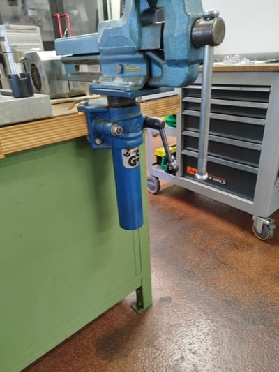 This image depicts a workshop environment in Bremerhaven, Germany. The foreground is dominated by a heavy-duty blue vise attached to the edge of a green workbench. The vise has a long silver handle and a blue cylindrical attachment hanging below it. Behind the workbench, a grey rolling tool cabinet with multiple drawers is visible, partially obscured by the vise and its attachment. The cabinet has various wheels, some with orange accents. The floor is a mottled reddish-brown. In the background, a glass-paneled door or window allows a glimpse into another area with what appears to be some sort of machinery or equipment, including red tubular structures and reflective surfaces. The overall scene suggests a functional, industrial or mechanical workshop setting.