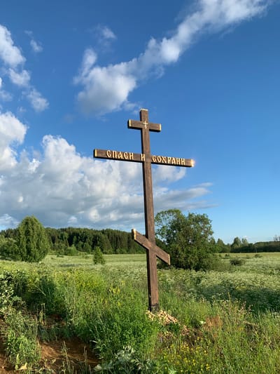 The image shows a wooden Orthodox cross in a field in the village of Svyatitsa, Russia. The cross is dark brown and has gold lettering on one of its beams. The lettering appears to be in Cyrillic script and reads "СПАСИ И СОХРАНИ" (Save and Protect). The cross is positioned in the foreground, and behind it is a vast, green field dotted with white and yellow wildflowers. A dense forest borders the field in the background, and the sky above is a clear blue with scattered white clouds. The lighting suggests it is daytime, possibly late afternoon due to the golden glow on the cross's lettering. There are no people visible in the image. The environment is natural and rural.