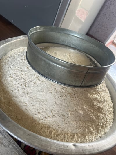 In Kara-Dıykan, Kyrgyzstan, a metal sieve rests atop a large metal bowl filled with white flour. The sieve, partially obscuring the flour within the bowl, also has some flour resting on its mesh. The bowl itself appears to be filled to a significant level with the fine powder. The scene is likely indoors, possibly in a kitchen or food preparation area, given the presence of kitchenware.