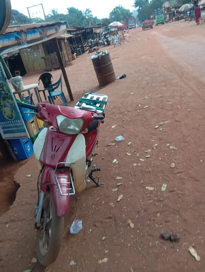 The scene depicts a rural or semi-urban roadside market area in Okpatu-Ukehe, Nigeria, under an overcast sky. In the immediate foreground, a red and beige scooter with a front metal rack and a rear green and white luggage rack is parked facing slightly right on a red-brown dirt verge. To the left of the scooter, a blue plastic crate is visible, along with a white plastic table holding a translucent container and a green bottle. Two plastic chairs, one blue and one black, are positioned around the table. A vertical sign with text and images, partially reading "CHIMDALU," stands nearby. The ground is consistently red-brown dirt, littered with scattered dry leaves, plastic debris, and visible footprints.

In the midground, running parallel to the left side of the dirt road, is a row of informal market stalls constructed with wooden frames and corrugated metal roofs. One stall displays a sign reading "TURU UGO LOTA." An old, rusty metal barrel, partially filled with refuse, stands on the dirt verge next to a sign labeled "P.O.S." The unpaved red-brown dirt road extends into the distance, showing tire tracks. Multiple vehicles are observed on the road, including a green small truck, a lighter-colored larger truck, and a red vehicle, all moving away from the viewer. Several individuals are present on or near the road; one person in a dark red garment is walking on the far right side, moving away. The background consists of dense green foliage, and overhead power lines are visible against the hazy sky.