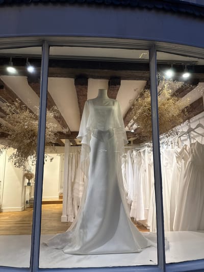 A store window displays a white wedding dress on a mannequin. The dress's design features ruffles around the neck area and a long, flowing train. Additional dresses are visible on racks inside the store. The interior architecture includes exposed wooden beams. Decorative dried flowers are arranged around the mannequin and along the beams. The window frame is painted blue.