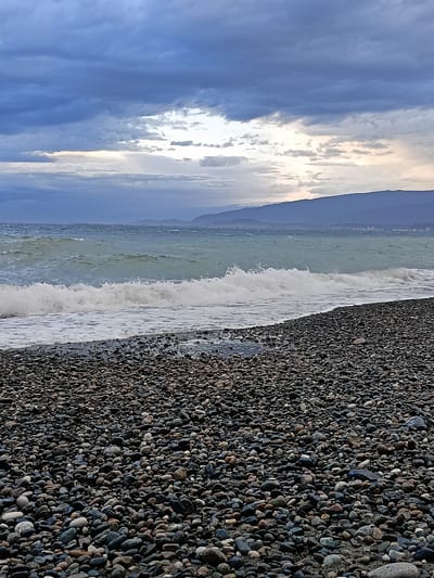 The image depicts a pebble beach in Gulrypsh, Abkhazia. The foreground is dominated by a dense expanse of smooth, rounded pebbles in shades of grey, black, and brown. A wave is breaking on the shore, creating a band of white foam separating the pebbles from the sea. The sea itself is choppy with a grey-green color. In the background, a coastline stretches towards a mountain range under a cloudy sky. The sky is predominantly grey with hints of lighter areas. The overall lighting suggests an overcast or pre-sunset time of day. There are no people or other visible signs of human activity.