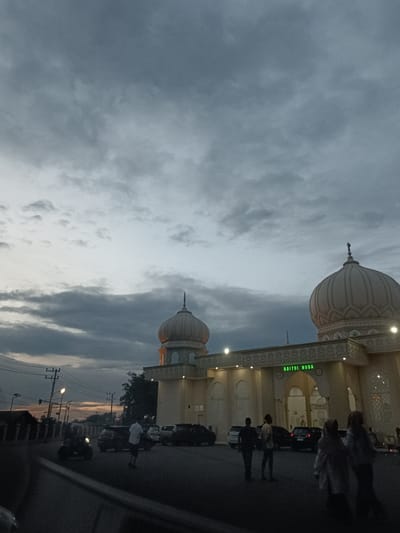 This media file captures a scene at dusk in front of a mosque named "BAITUL HUDA" in Mee Tanjong Usi, Indonesia. The sky is filled with dramatic, dark gray clouds, suggesting overcast or stormy weather. The mosque, an imposing structure with prominent domes and intricate architectural details, is illuminated by artificial lights, creating a warm glow against the dimming sky.
Several individuals are visible in the foreground and near the mosque entrance. Some are walking, while others appear to be standing and observing. A few cars are parked in front of the mosque, and a motorcycle is also present on the road. Power lines and streetlights are visible in the background, indicating an urban or semi-urban setting.
The overall mood of the image is serene yet somewhat dramatic due to the cloudy sky. The time of day is clearly indicated by the transition from daylight to evening, with the lights of the mosque and streetlights beginning to dominate the illumination. The presence of people suggests that the mosque is a place of gathering or prayer, and the activity appears to be the end of the day's activities. The text "BAITUL HUDA" is prominently displayed in green neon letters above the mosque's entrance.