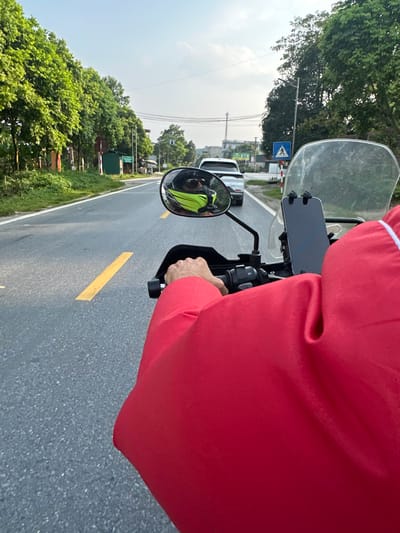 The image provides a first-person perspective from a person riding a motorbike along a paved road in Vị Xuyên Commune, Vietnam, during clear daylight hours. The rider, wearing a red long-sleeved top, has their right hand gripping the motorbike's throttle. A smartphone is mounted on the handlebars, situated just beneath a clear windshield. The rider's reflection, clad in a bright yellow or green garment, is visible in the left rearview mirror.
The asphalt road features a prominent solid yellow line on the left side and a white dashed line on the right, guiding traffic flow. Both sides of the road are bordered by abundant green trees and lush foliage, indicating a verdant, possibly rural or semi-urban setting. In the distance, several cars can be seen on the road.
Notable roadside features include a blue square sign displaying a white pedestrian crossing symbol on the left side of the road. Further down, a red and white striped barrier, characteristic of a railway crossing, is visible, indicating an upcoming intersection with a train track. Utility poles and overhead wires are also part of the landscape. The bright, clear sky suggests sunny weather, offering good visibility for travel. The scene captures a moment of peaceful transit through the Vietnamese countryside.