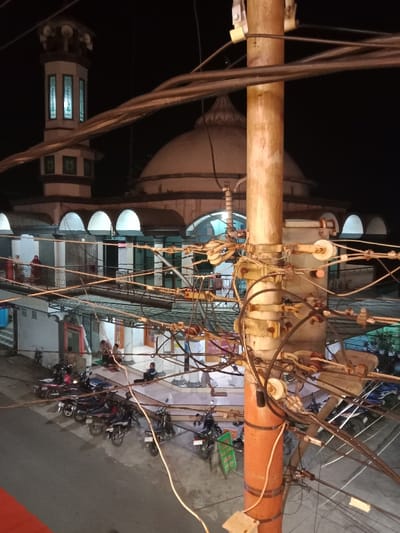 The image depicts a nighttime urban scene in Pandean, Indonesia. A utility pole, heavily laden with numerous electrical and communication wires and insulators, dominates the right foreground. In the background, a mosque with a prominent central dome and a multi-tiered minaret is visible, illuminated internally. Below the mosque's arched ground level, approximately eight motorcycles are parked on a paved surface. Several individuals are present: two figures are on an elevated balcony, one identifiable as a woman in a red head covering, while five to seven individuals are seated on mats near the mosque entrance at ground level. Additional indistinct figures are present within the illuminated architectural arches of the mosque. The environment is artificially lit against a dark night sky.