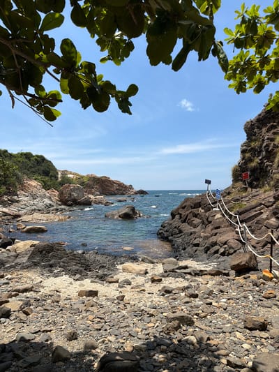 The image depicts a coastal scene with a rocky beach and clear blue water. Lush green foliage frames the top of the image, suggesting a tropical or subtropical environment. The beach is covered with various sizes of stones and pebbles, with a section of lighter colored sand in the middle ground. Dark, rugged rock formations line the right side of the scene, with a white rope railing guiding a path along the edge. Several blue and white signs are visible attached to poles along the railing, and a red sign is affixed to the rock face further up. The water is calm, with small waves lapping at the shore. In the distance, more rocky hills covered in vegetation rise from the sea, and a few small boats can be seen on the water. The sky is a bright blue with a few scattered white clouds. The overall impression is of a natural, somewhat rugged coastline. The location is identified as Phú Lương, Vietnam.