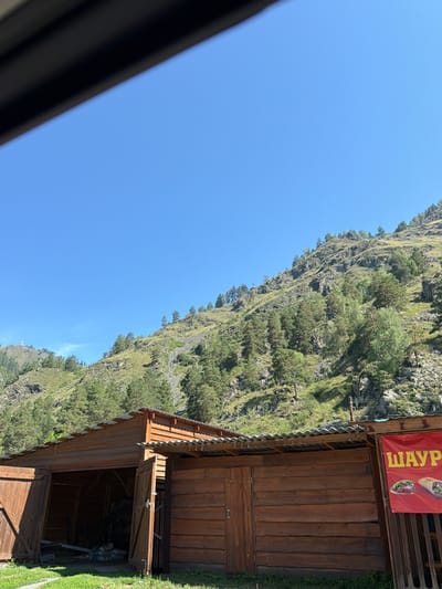 In the city of Uznezya, Russia, a scene unfolds beneath a clear blue sky, with a prominent mountain slope covered in lush green trees and rocky outcrops dominating the upper portion of the frame. The mountain is a vibrant landscape of mixed coniferous and deciduous trees, with patches of exposed rock and scree. A white structure, possibly a monument or a small building, is visible on a distant peak to the left. In the foreground, a wooden structure resembling a garage or workshop with an open bay stands. Adjacent to it, to the right, is another wooden building with a red banner that reads "ШАУРМА" in large yellow Cyrillic letters. The banner also features images of shawarma. The wooden structures appear rustic, built from logs or planks, and have corrugated metal roofs. Green grass grows in front of the buildings. The overall impression is a rural or semi-rural setting with a food vendor operating near a mountainous natural landscape.
