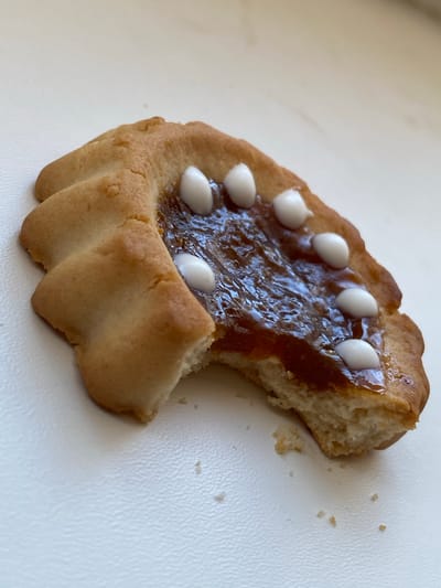 A close-up shot shows a partially eaten cookie with amber-colored jam and white candy dots on a white textured surface. The cookie is golden brown and has a bite taken out of its lower right side. The jam is glossy and covers a portion of the cookie's surface, where seven small, oval-shaped white candy dots are arranged in a curved line. The background is out of focus. The image does not contain information about the city of Bobrovo, Russia.