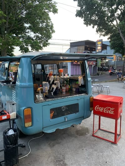 A light blue vintage Volkswagen Type 2 van, modified as a mobile beverage vendor, is positioned on a paved surface in Landungsari, Indonesia. The van's rear hatch is open, exposing a counter equipped with an assortment of clear and tinted plastic and glass bottles, shakers, cups, and an orange-bristled brush. A red Coca-Cola branded cooler on a red metal stand stands beside the van's right rear. To the van's left, a black trash receptacle is situated next to a small black table holding white containers and an orange tray. White electrical cabling extends from the van. In the mid-ground, multiple white metal tables and matching stools are arranged for customer seating, with one individual in a striped shirt occupying a stool. The background reveals a paved street with parked motorcycles and one moving scooter. Commercial buildings with assorted signage are visible beyond the street, framed by large deciduous trees under an evening sky.