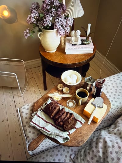 This indoor still life depicts a cozy breakfast or snack setup within a warm, inviting setting. The scene is captured from an overhead perspective, looking down onto a light wooden floor.

On the right side of the frame, a sofa or armchair with light-colored, polka-dotted upholstery holds a paddle-shaped wooden serving board. On this board, a meal is arranged: slices of dark bread rest on a striped linen towel, alongside a block of yellow cheese with a metal cheese slicer. A small jar of red jam with a spoon is visible next to the cheese. A light-colored mug contains coffee, and a small white bowl holds three white eggs. Complementing these are a tiny bowl of white salt, a small container with butter and a wooden spreading knife, and a small metal pitcher, possibly for milk or cream.

To the left, a round wooden side table holds a light-colored ceramic pitcher brimming with vibrant purple lilac flowers. A white, pleated lampshade stands nearby. Also on the table, a stack of books acts as a base for a small white ceramic figurine, appearing to be a cherub-like figure, which holds a slender white candle.

At the bottom of the frame, on the wooden floorboards, a light-colored metal rack, possibly for magazines or blankets, is visible. The lighting suggests daytime, contributing to the serene and comfortable atmosphere.

The visible text on one of the white book spines reads "NYCKELN till HEMMET," which is Swedish for "The Key to the Home."