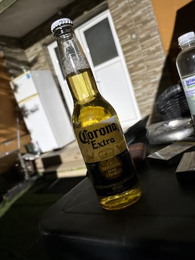 The image, captured in Dolna banya, Bulgaria, presents a close-up of a Corona Extra beer bottle lying horizontally across the frame. The amber-colored bottle shows visible condensation and foam concentrated around its neck, suggesting it is cold and freshly opened or agitated. The bottle's cap remains secured.

In the upper left background, a clear plastic water bottle with a white cap is partially visible, bearing text on its label that reads "DEVINE Mineral Vitamin." The immediate foreground and right side of the image feature a dark, unidentifiable surface, possibly a table or counter. Indistinct metallic or plastic items are also discernible in the background.

Behind the bottles, a light-colored structure, likely a window or door frame with a dark pane, provides a backdrop, indicating an indoor setting. The overall lighting is subdued, suggesting the image was taken in the evening or at night.

Visible text on the Corona Extra bottle includes the brand name "Corona Extra," the slogan "LA CERVEZA MAS FINA" (The Finest Beer), and production details such as "CERVECERIA MODELO S. DE R.L. DE C.V.," "MEXICO," and "REG. SSA. No. 7417." The water bottle's label displays "DEVINE Mineral Vitam[in]." No individuals are present, and no specific activity is taking place beyond the arrangement of the beverages.