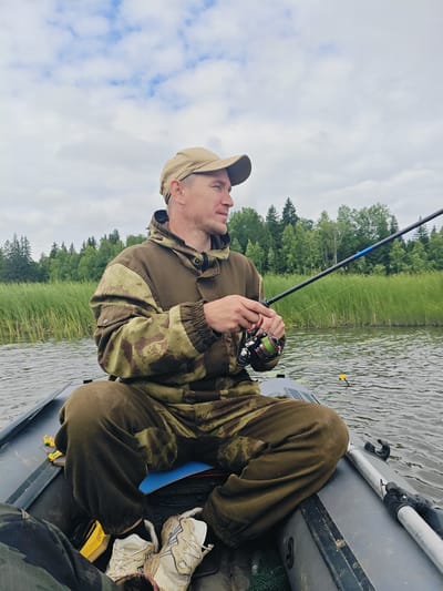 A man is fishing from an inflatable boat on a lake. He is wearing a camouflage jacket and a beige baseball cap. He is holding a fishing rod and reel. The boat is in a body of water with reeds and trees on the shore. The sky is cloudy. The scene appears to be in Detskii Dom (Bashenino), Russia.