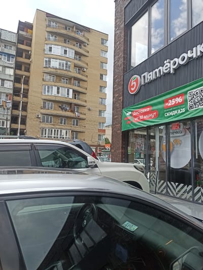 The image shows a street scene in Atabe, Georgia, with a focus on a "Pyaterochka" grocery store. The store has a prominent sign with the "Pyaterochka" logo, a red circle with a white "5" inside. A green banner is displayed, advertising "Delivery from 30 minutes" and a "-25% discount for the first order with code SKIDKA25". The store entrance has large windows, with some of the interior visible, including shelves with products and possibly some advertising posters.
In front of the store, several cars are parked. The perspective is from inside one of these cars, looking out. The interior of the car is visible, including the dashboard, steering wheel, and part of the passenger side. A sticker or decal is visible on the windshield.
Behind the store, a tall, multi-story building with a distinctive architectural style is visible. The building is made of light-colored brick or concrete and features a curved, cantilevered section with many windows. Laundry is hanging from some of the balconies on this building. The sky is overcast, suggesting a cloudy day. The overall impression is an urban street view, likely during daylight hours.