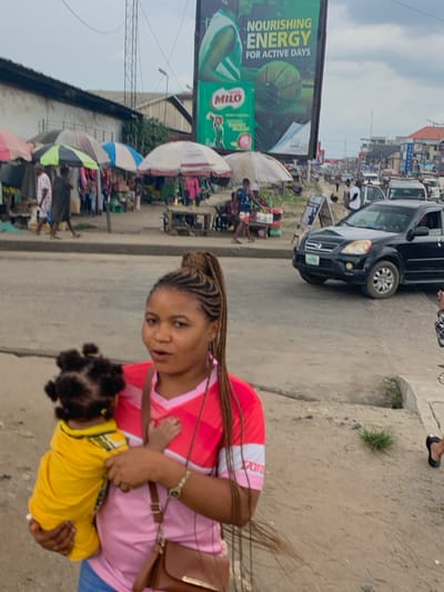This image depicts a bustling urban street scene in Aba, Abia, Nigeria. In the immediate foreground, a woman, appearing to be in her twenties or thirties, stands holding a small child. The woman has light brown skin, her hair is styled in long, intricate cornrow braids pulled into a high ponytail, and she wears a pink and red short-sleeved shirt with blue jeans. A brown shoulder bag is slung across her body. She looks directly towards the viewer with her mouth slightly open. The child she holds is clad in a yellow shirt, with their back to the camera, revealing dark hair styled in multiple small buns or knots.
Behind them, a paved road stretches diagonally, showing active traffic. A black SUV, possibly a Honda CR-V, is visible moving right-to-left in the mid-ground. Along the opposite side of the road, a vibrant market scene unfolds with numerous vendor stalls shaded by various large umbrellas in colors such as white, blue, green, and striped patterns. People are seen walking and standing among these stalls, indicating commerce. Further down the road, more vehicles and distant structures are visible under a light, overcast sky. A prominent feature in the background is a large billboard advertising "MILO" (Nestlé), displaying text that reads "NOURISHING ENERGY FOR ACTIVE DAYS" alongside an image related to sports. A tall metal mast is also present in the distance. The ground where the woman stands is an unpaved, sandy-colored shoulder adjacent to the asphalt road.