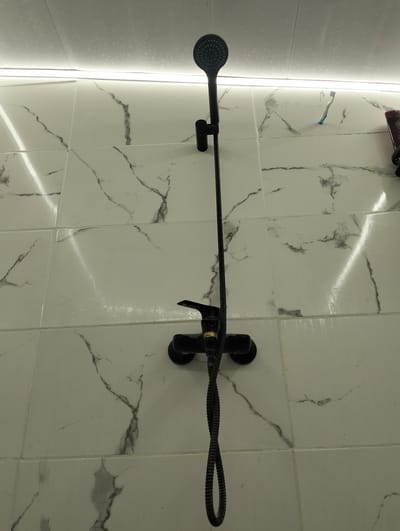 A black shower head and faucet are mounted on a white marble-patterned tiled wall. A strip of LED lighting is installed along the top of the wall. A toothbrush is visible on the wall to the right of the shower head. The scene appears to be a bathroom in an apartment in Șoldănești, Moldova.