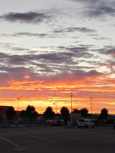 The image depicts a sunrise over a parking lot in Achern, Germany. The sky is filled with vibrant hues of orange, pink, and red, with darker gray clouds interspersed. In the foreground, an empty asphalt parking lot is visible, marked with white lines. Several streetlights stand tall throughout the lot. In the middle ground, a white van is parked. Behind the parking lot, there are trees, fences, and buildings. The overall scene suggests an early morning setting.