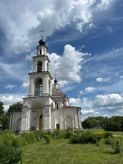 The image shows a white church with a bell tower against a bright blue sky with dramatic clouds. The church features arched windows and doorways, and the bell tower has multiple tiers of arches. A cross adorns the top of the bell tower and another dome. To the right of the church, there is a well-maintained grassy area with lush green bushes and trees. In the background, more trees and a hint of a fence are visible. The scene is illuminated by bright sunlight, suggesting it is daytime. The sky indicates it might be a partly cloudy day, with large, white clouds scattered across the blue. The overall impression is one of a peaceful and well-kept religious site in a rural or park-like setting. There is no visible text.