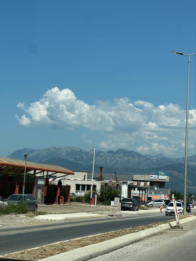 The image depicts a street scene in Bratešići, Montenegro, on a clear day with a bright blue sky and scattered white clouds. In the foreground, a paved road is visible, bordered by a construction zone with loose gravel and concrete barriers. Several cars are parked or driving on the road. To the left of the road, there is a car wash facility with an orange-topped structure. Behind the car wash, a low-rise building with a light-colored facade and windows is visible. Further back, a larger building with signage that includes "beko" and "BESIX SOLE CASA" is present. In the background, a mountain range with rugged peaks stretches across the horizon. A tall street lamp stands on the right side of the image. The overall scene suggests a commercial or industrial area on the outskirts of a town, with a backdrop of natural mountainous terrain.