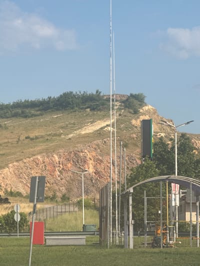 This is an outdoor, daytime, medium shot photograph taken in the vicinity of Polsko Padarevo, Bulgaria. The scene depicts a scrubby, hilly landscape with a large, orange-brown rocky incline dominating the background. The foreground features a grassy area with a road and some industrial-looking structures.
To the left, a gray pole with two circular signs is visible. Next to it is a tall, red rectangular object. Further right, a chain-link fence encloses an area with gas piping and a yellow ladder. A large, dark, rectangular digital billboard is positioned on a pole behind the fence and trees. Several tall, thin poles, possibly for lighting or antennas, are scattered throughout the scene. The sky is light blue with scattered white clouds.