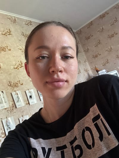 A selfie of a person with light skin, light eyes, and dark hair, wearing a black t-shirt with a white printed design. The person is indoors with a wall behind them covered with a patterned wallpaper and a series of small framed pictures hanging on the wall. The ceiling is visible at the top left corner. There is no information about Ogryzkovo, Russia available in the image.