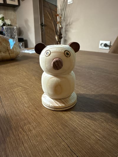 A close-up, eye-level, slightly angled shot shows a small, wooden, carved bear figurine on a wooden table. The bear is light-colored wood, with dark brown, round ears and a dark brown, triangular nose. The eyes are circular with dark pupils. The bear's body is made of two stacked spheres, and it sits on a circular wooden base. The table has a warm, brown, wood grain pattern. In the blurred background, to the left, is a loaf of bread in plastic packaging, and some white flowers in a glass vase. To the right, a wall is visible with a white electrical outlet. The scene is indoors, likely in a home or a shop. The specific location of Goliam Dol, Bulgaria, is not evident from the visual cues in the image.