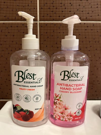 Two bottles of Blest Essentials Antibacterial Hand Soap are positioned side-by-side on a white surface against a tiled brown background. The bottle on the left is clear and contains a light-colored liquid, labeled "FRUITY FRESH" with an illustration of various berries. The bottle on the right is clear and contains pink liquid, labeled "CHERRY BLOSSOM" with an illustration of cherry blossoms. Both bottles have white pump dispensers. The scene is indoors, with the tiled wall suggesting a bathroom or kitchen environment. There are no people or animals present in the image. The lighting appears to be artificial. The image does not provide any specific information about its location in Quezon City, Philippines, or any identifiable landmarks.